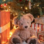 Teddy bear is sitting under Christmas tree.